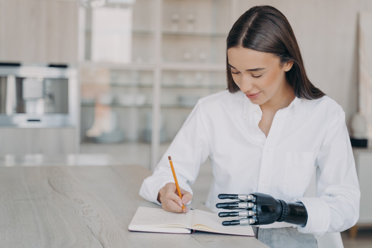 Modern pretty girl with disability writing in notebook, sitting at table at home. Disabled young female makes notes, writes to-do list, planning goals in notepad, using bionic prosthetic arm.